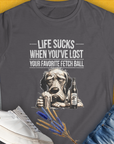 Charcoal womens t-shirt, "Life Sucks When You've Lost Your Favorite Fetch Ball", Volume 5 from the popular series of the same name. Funny drawing of a depressed dog face. Next to the dog is a full glass and a bottle with a label that says "Chicken Soup with Whipped Cream, for Sad Dog Days". Designed by Dog Gang.