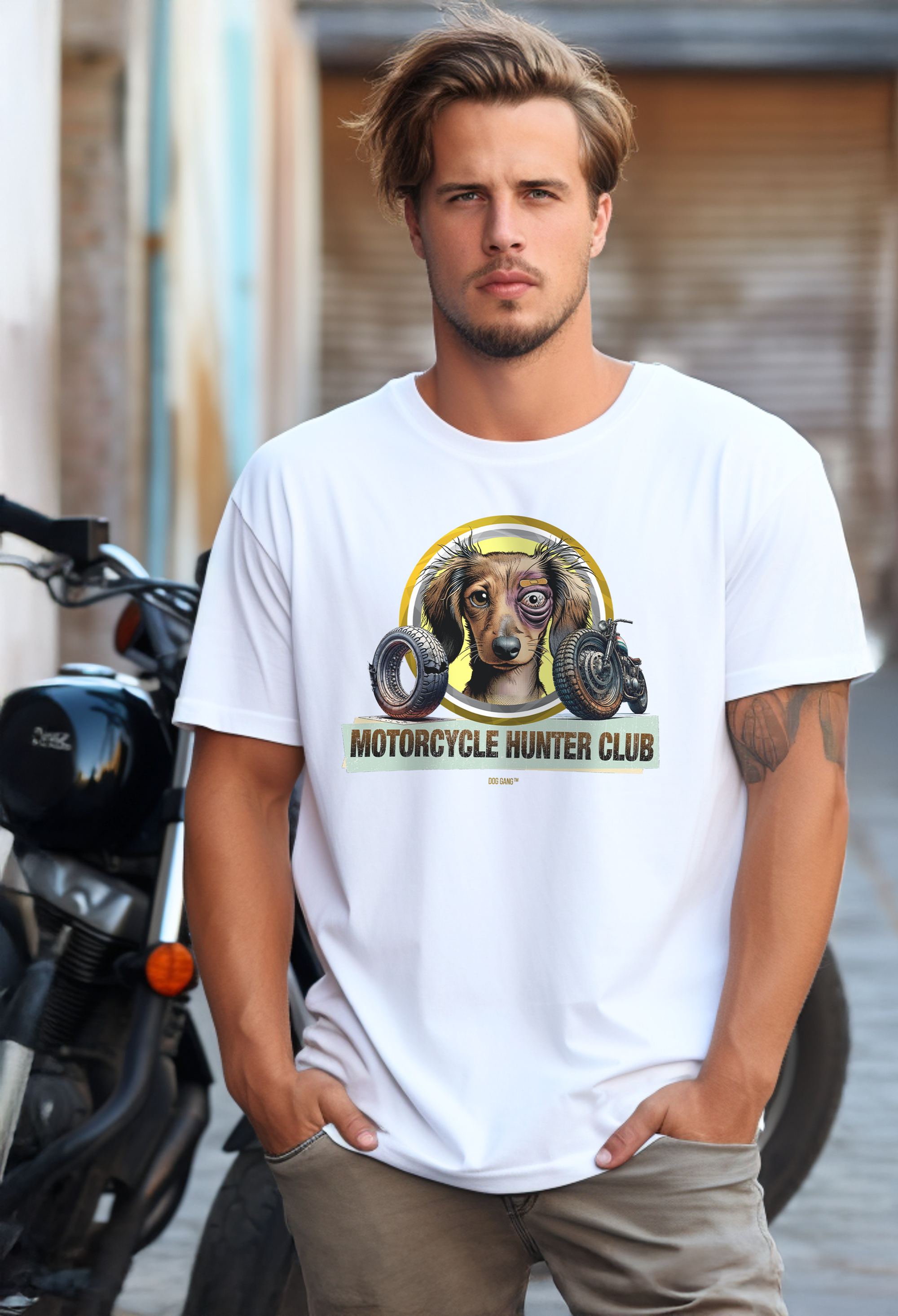 A man with short hair stands on a street in front of a motorcycle. He is wearing a white Dog Gang T-shirt with a Motorcycle Hunter Club motif showing a slightly disheveled but happy dog face with a black eye and bitten motorcycle parts.