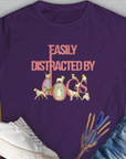A purple, semi-fitted womens t-shirt from the Easily Distracted By Dogs Series, Volume 2, features bold Easily Distracted by Dogs text in pink and multicolored letters. The word dogs is creatively shaped with dog illustrations: a Dalmatian, a Golden Retriever, a Pug, a Poodle, Dachshund, English Bulldog, Chihuahua, Husky and more. Designed in Switzerland by Dog Gang.