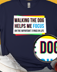 Navy blue shirt featuring a funny dog design with a quote: Walking the dog helps me focus on the important things in life, alongside shoes and a ball. Designed by Dog Gang.