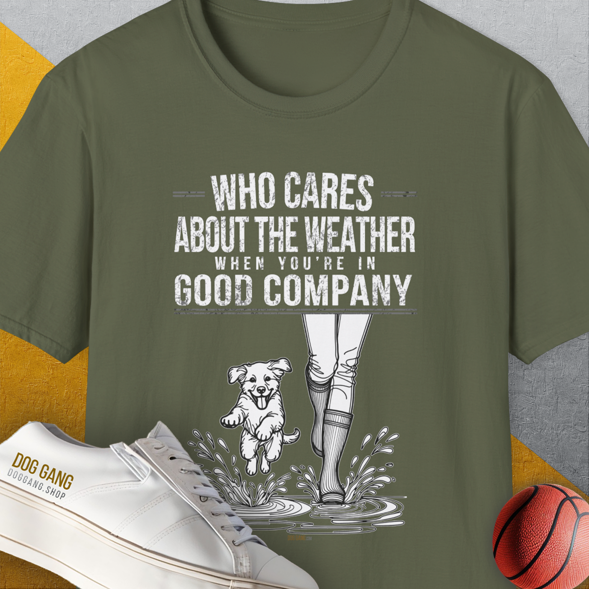 Military green tee with a dog and person in rain boots. Text: Who Cares About The Weather When Youre In Good Company, Volume 2. Perfect for any dog person. Designed by Dog Gang.