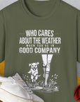Military green tee with a dog and person in rain boots. Text: Who Cares About The Weather When Youre In Good Company, Volume 2. Perfect for any dog person. Designed by Dog Gang.