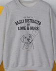 The sweatshirt titled My Dog Is Easily Distracted By - Volume 3: Love And Hugs showcases the phrase in bold black lettering on a gray heavy-blend crewneck. The design includes a minimalist dog face and heart line drawing, enhancing its charm. Made from ethically sourced cotton, this piece boasts textured surfaces in gray and yellow. Shirtdesign by Dog Gang adds a unique touch to this stylish garment.
