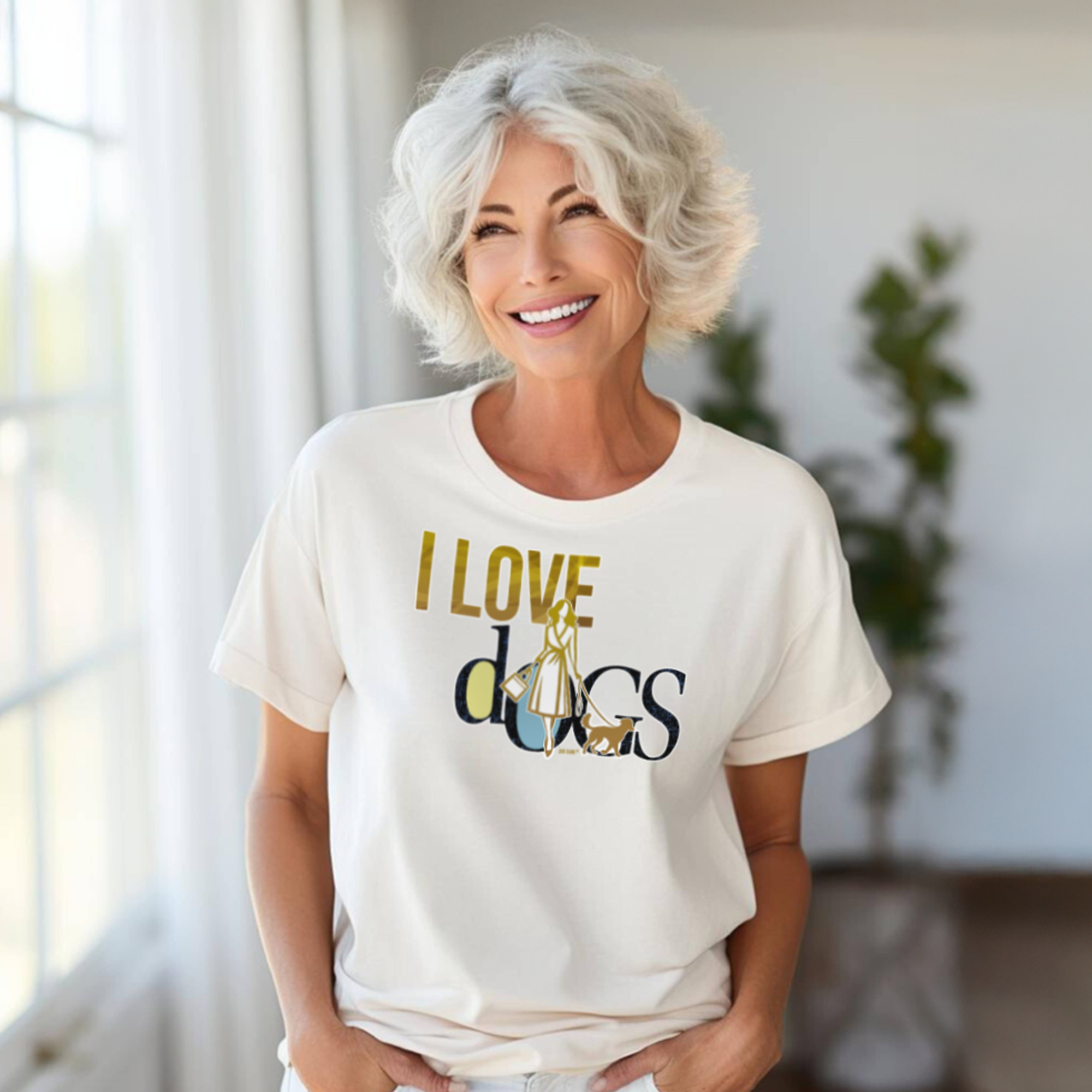 A woman with short gray hair is smiling and wearing a white T-shirt that says I LOVE dogs in large letters. She is standing in a bright room with two blurred green plants in the background.