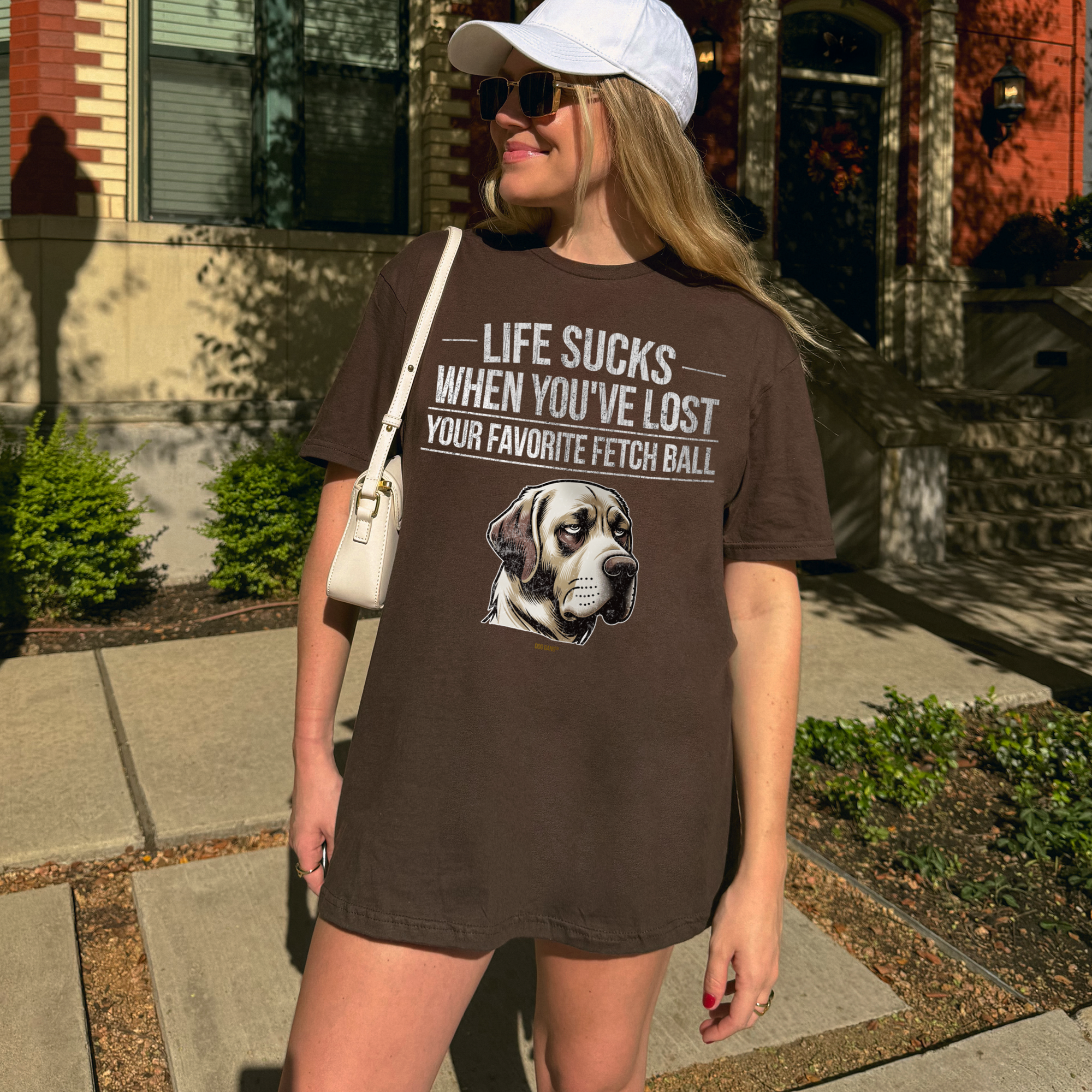 A person stands on a sidewalk wearing a brown T-shirt with a dog image and the text Life Sucks When Youve Lost Your Favorite Fetch Ball. They have sunglasses, a white cap, a white shoulder bag, and there is a building in the background.
