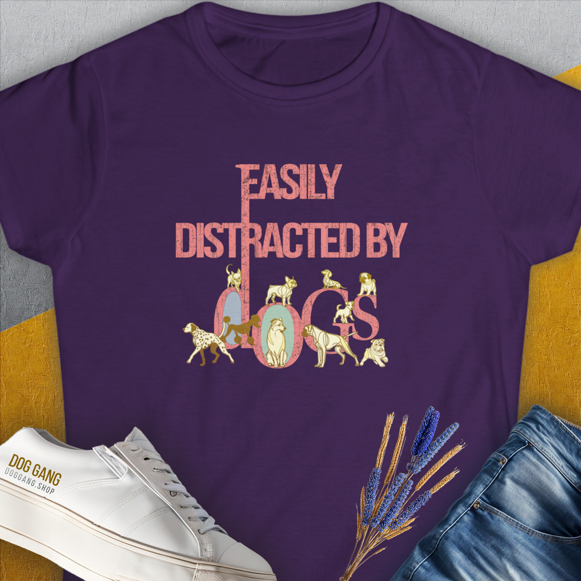 A purple, semi-fitted womens t-shirt from the Easily Distracted By Dogs Series, Volume 2, features bold Easily Distracted by Dogs text in pink and multicolored letters. The word dogs is creatively shaped with dog illustrations: a Dalmatian, a Golden Retriever, a Pug, a Poodle, Dachshund, English Bulldog, Chihuahua, Husky and more. Designed in Switzerland by Dog Gang.