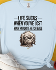 This light blue unisex T-shirt features a sad dog illustration with the quote: Life Sucks When Youve Lost Your Favorite Fetch Ball, Volume 2. Perfect for a dog lover. Designed by Dog Gang.
