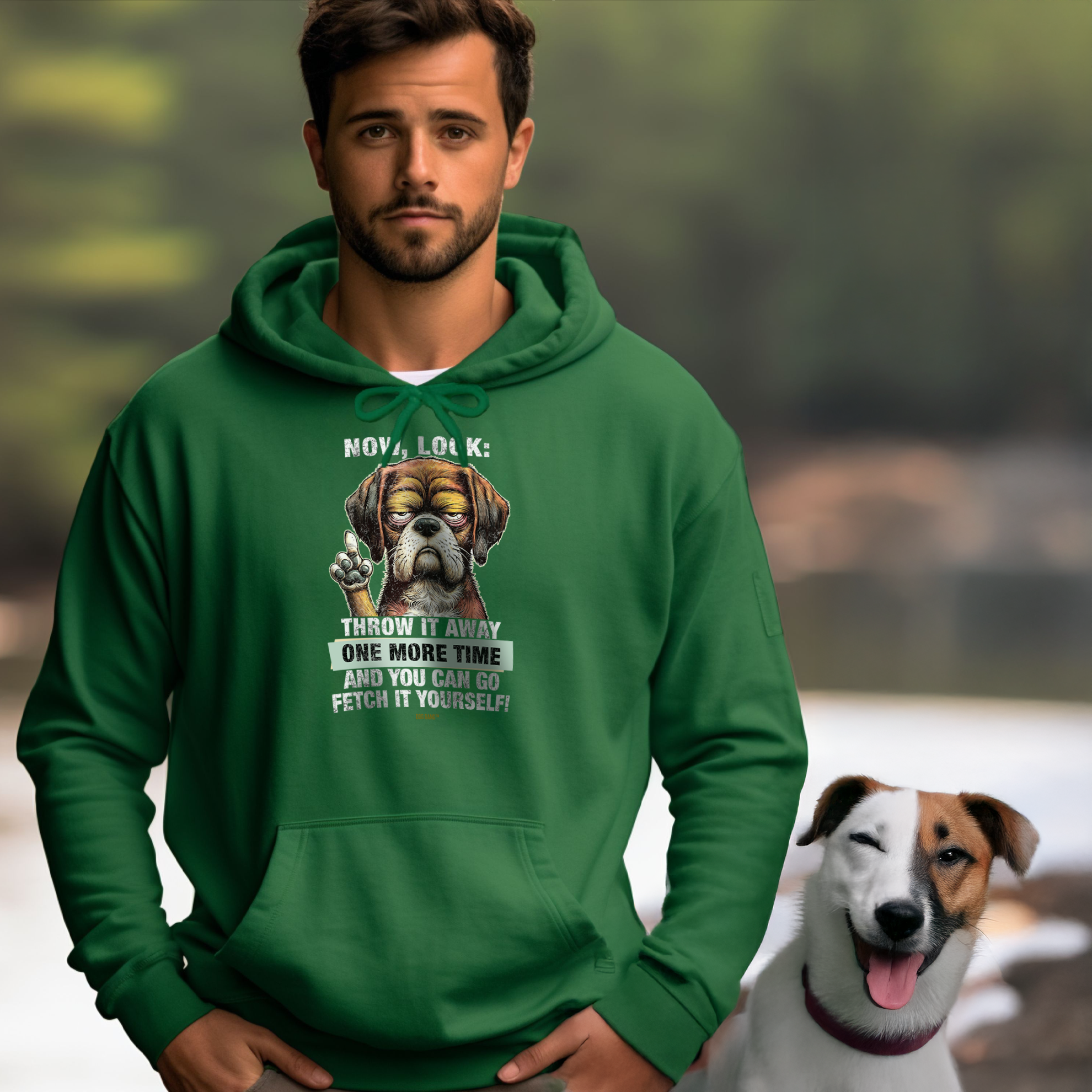 A man wearing a green hoodie with a graphic of a grumpy dog and humorous text stands outdoors. A happy dog sits beside him, winking with its tongue out. The background features blurred outdoor scenery.