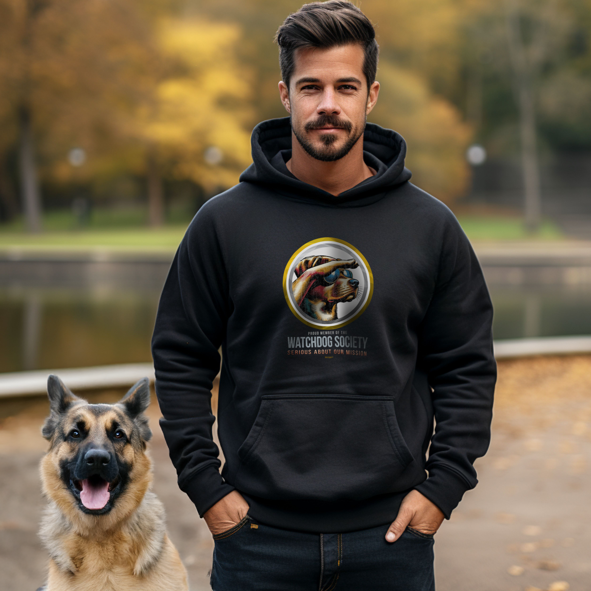 A man wearing a black hoodie with a Watchdog Society logo stands next to a German Shepherd. They are outside near a body of water with trees in the background. The scene is set in an autumnal environment.
