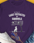 The My Dog Is Easily Distracted By - Volume 1: Squirrels womens T-shirt in purple showcases a funny dog design with a playful pup watching squirrels and the quote my dog is easily distracted by squirrels. Designed by Dog Gang.
