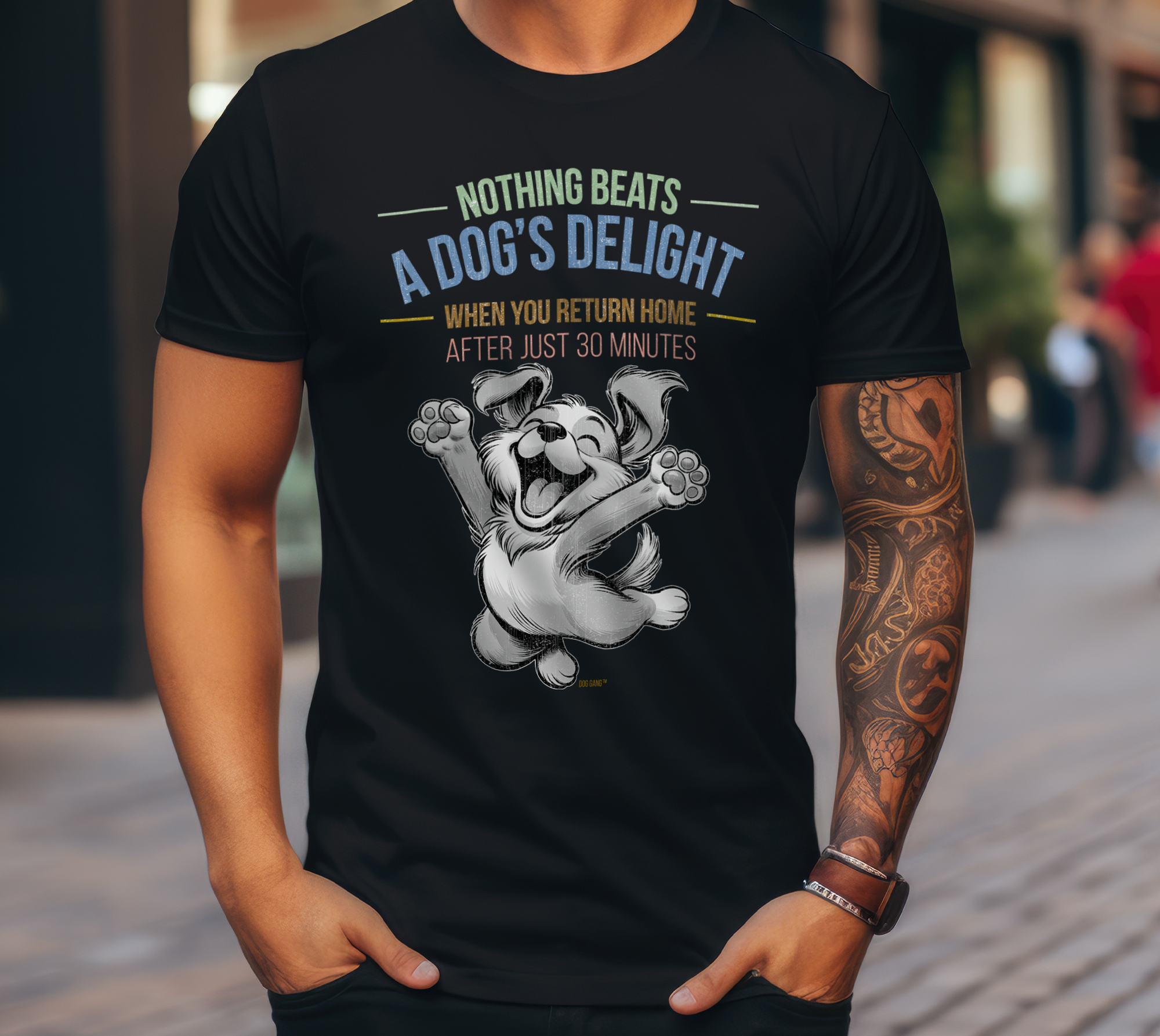 A person with a tattooed arm wears a black Dog Gang t-shirt with a cartoon dog jumping happily. The shirt reads: "Nothing beats the joy of a dog when you get home after only 30 minutes." A blurry street scene can be seen in the background.